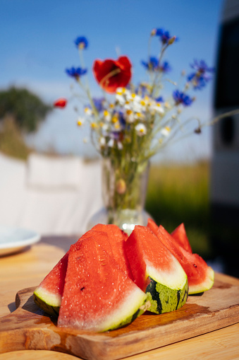 Sliced watermelon laying on a wooden board on a table outside, under the sun with a vase of wildflowers in the background. The image captures the refreshing taste of summer and the natural beauty of the surrounding wildflowers.