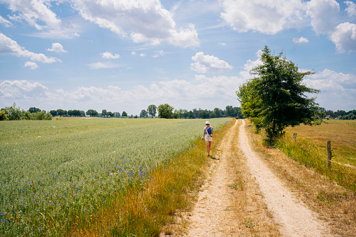 A woman is walking on a dirt road surrounded by an agricultural field and trees in the background. The image captures the simplicity and beauty of the rural landscape on a sunny day with blue sky and white clouds.