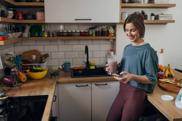 eine lächelnde frau trinkt einen milchshake und benutzt ein handy, während sie in der küche steht - juicer food and drink healthy eating sport stock-fotos und bilder