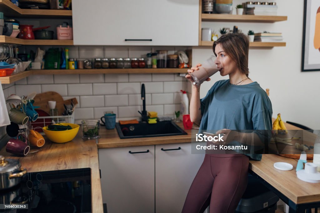 An athletic woman drinks a milkshake she made in the kitchen and surfs the Internet Fit woman drinking healthy fruit milkshake for breakfast while holding mobile phone in hand Milkshake Stock Photo
