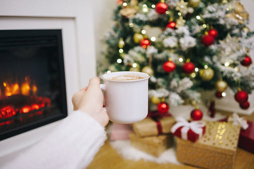 POV - woman holding a cup with hot chocolate in front of the decorated Christmas tree and fireplace. Cozy Christmas at home concept. Relaxing holidays alone.
