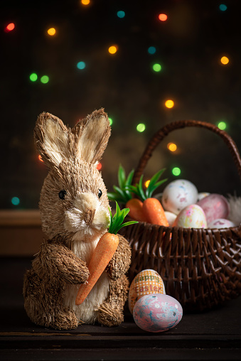 Easter bunny with a carrot in his hands next to a basket with Easter eggs in front of a lighted window with blurred festive background