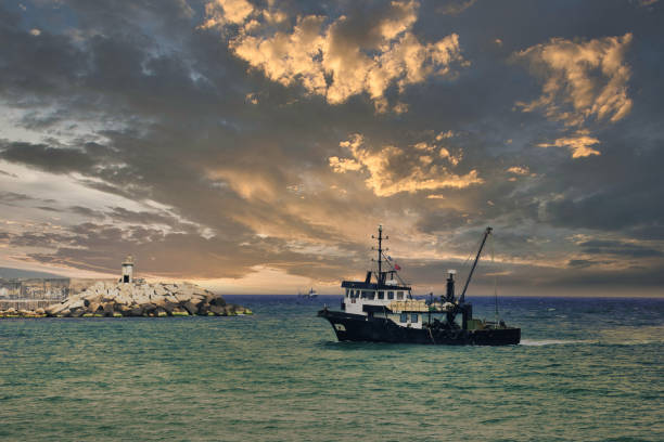 vista de un barco de pesca que regresa del mar a tierra al atardecer y una vista del pequeño puerto, el faro y el hermoso cielo dramático. - retro fish day sunset sunlight fotografías e imágenes de stock