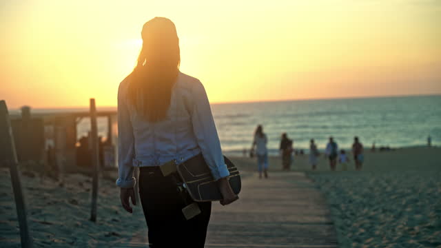 Woman with brown hair looking at the view,walking to the beach while carrying a skateboard underneath her arm,during sunset