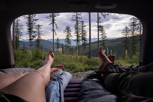 A couple enjoys the view while lying in the back of a SUV. They watch the end of a sunset overlooking a valley filled with pine trees.
