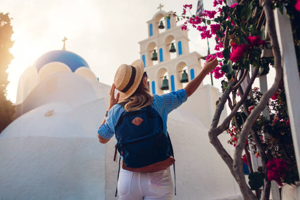Santorini tourist with backpack walks by church with blue dome and blooming bougainvillea flowers in Akrotiri stock photo