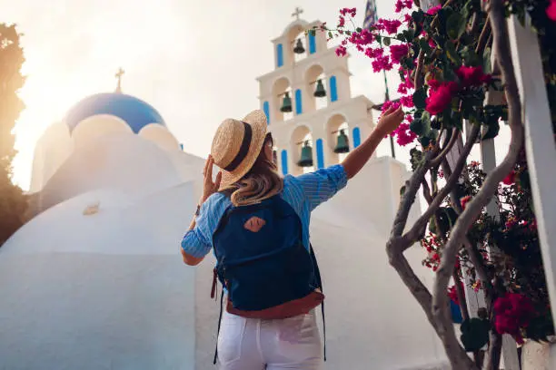 Santorini tourist with backpack walks by church with blue dome and blooming bougainvillea flowers in Akrotiri at sunset. Summer vacation