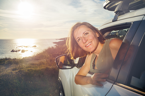 Young woman sticking out head and hands of the car driving door window smiling looking back over ocean sunset