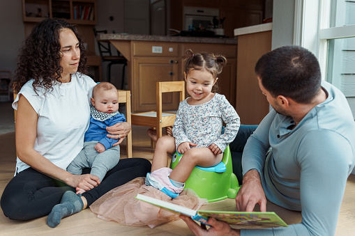 An adorable little girl who is practicing using the potty smiles as her dad reads her a picture book. The toddler's mom is holding her baby brother and sitting on the floor next to her.