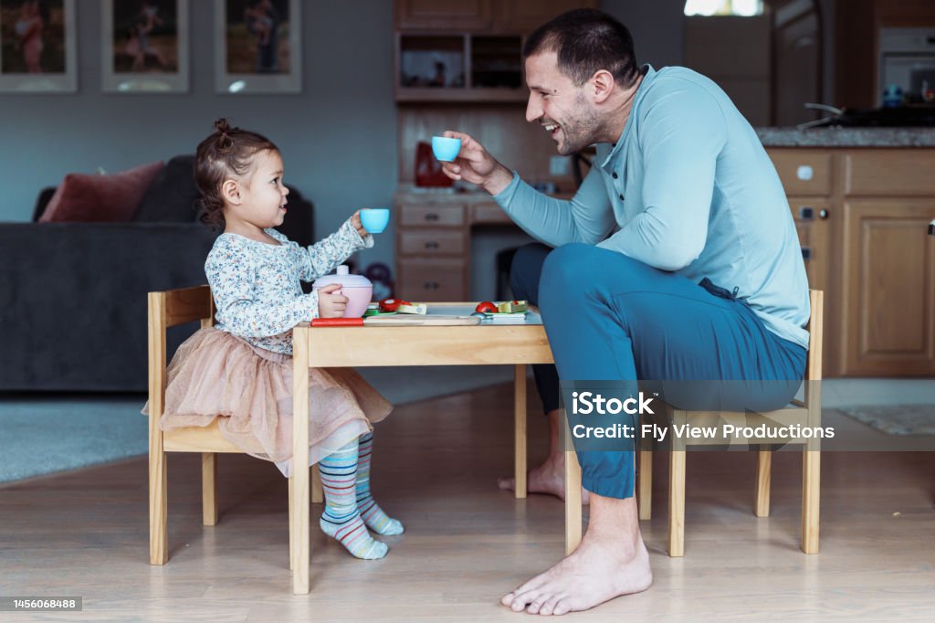 Toddler girl and dad toast while having tea party Profile view of a dad and his toddler daughter sitting at a child size table and doing a celebratory toast with toy teacups while playing together at home. Family Stock Photo