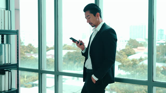 Asian young adult businessman is leaning against mirror wall and checking social media with smartphone during break.