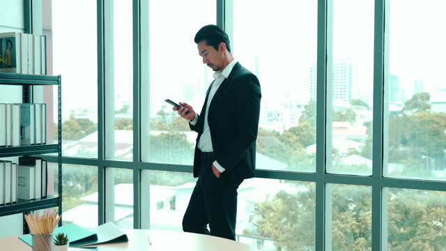 Asian young adult businessman is watching online news on his smartphone while leaning against mirror wall in the office.