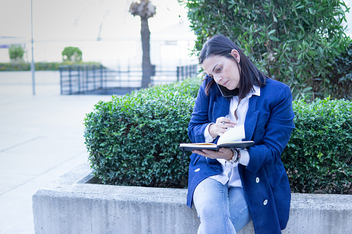 Beautiful Caucasian businesswoman calling phone and wrting in a notebook outside an office building. She is wearing a casual blue jacket.