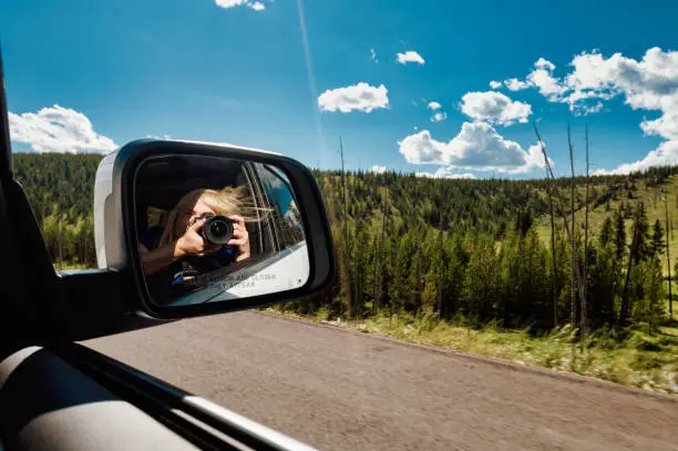 Photo of Woman on a road trip takes a photo with a camera out of a car window reflected in side mirrors of stunning natural landscape