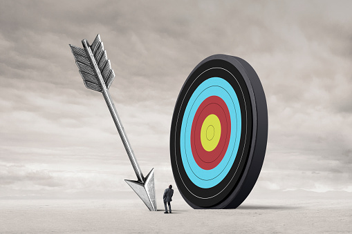 A man looks up at a large arrow stuck in the ground that has missed its target.