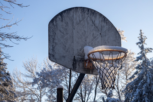 This image shows a close-up abstract low angle view of a grungy old outdoor basketball hoop and backboard, containing snow from a recent blizzard with back lit sunlight.