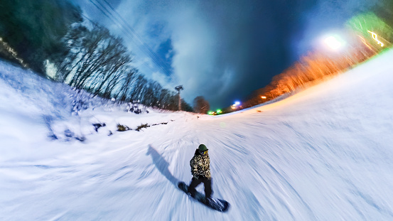 A wide angle self photo of a snowboarder riding down a ski slope at night time. There is blurred motion.