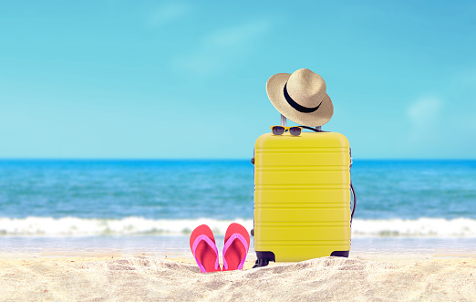 Yellow luggage with hat and red flip-flop on sandy beach under blue sky background.