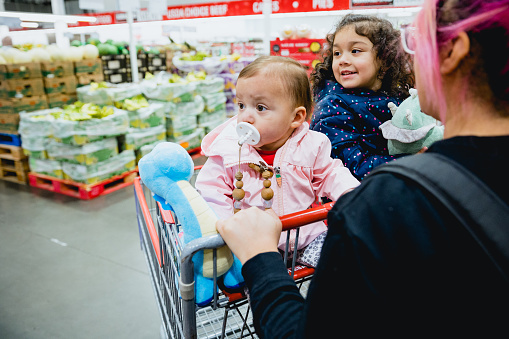 Lifestyle photos of a mom with two toddler children grocery shopping inside a supermarket. The two children are in the shopping cart as the mom adds food items to the cart.