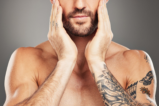 Beauty, skincare and body with a man model in studio on a gray background with an arm tattoo for wellness. Health, luxury and skin with a male touching his face for cleaning or natural bodycare