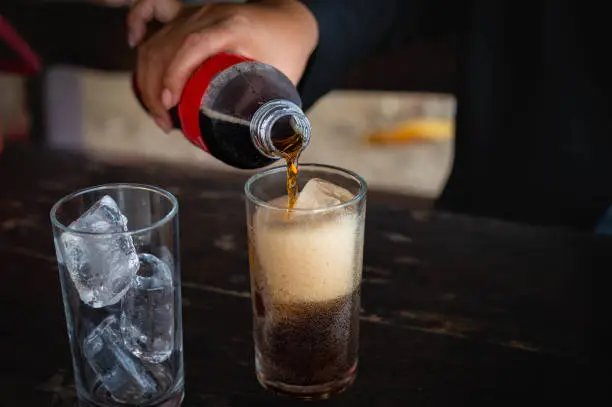 Woman holding pouring cold cola soft drink soda, sparkling water with ice sweet sugar from bottle into glass in her hand. Health care, healthy diet lifestyle concept.