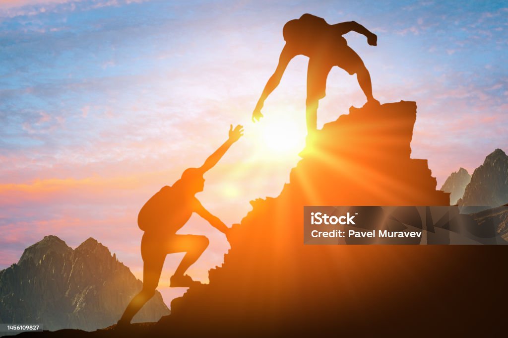 Der Mensch gibt helfende Hand. Silhouetten von Menschen, die bei Sonnenuntergang auf den Berg klettern. Hilfe- und Assistenzkonzept. Silhouetten von zwei Menschen, die auf den Berg klettern und helfen. - Lizenzfrei Unterstützung Stock-Foto