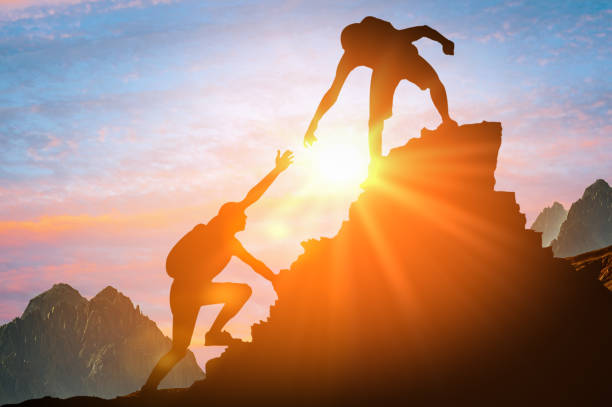 Man is giving helping hand. Silhouettes of people climbing on mountain at sunset. Help and assistance concept. Silhouettes of two people climbing on mountain and helping. Help and assistance concept. Silhouettes of two people climbing on mountain thanks to mutual assistance and teamwork and partnership. business success and teamwork concept in company hulp stock pictures, royalty-free photos & images