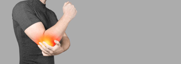 グレイの背景に肘の痛みを持つ男性。慢性関節リウマチを患っている男性。理学療法による共同事故。バナー。コピースペース - pain elbow physical therapy inflammation ストックフォトと画像