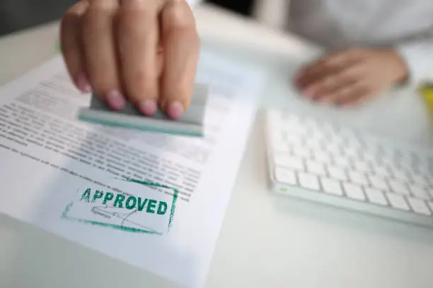 Photo of Closeup of a person hand stamping green approved seal on text approved document on table