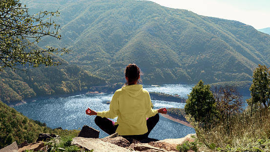 Young woman practicing yoga and meditating on nature. Back view girl relaxing on background of amazing landscape of mountain lake. Mindfulness, mental health, relaxation, vitality energy
