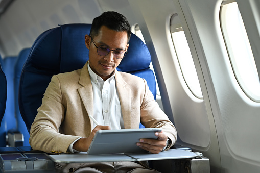 Millennial male entrepreneur working on digital tablet sitting near window in an airplane.