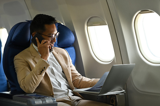 Professional businessman in formal suit talking on mobile phone and using laptop while sitting in airplane cabin near window.