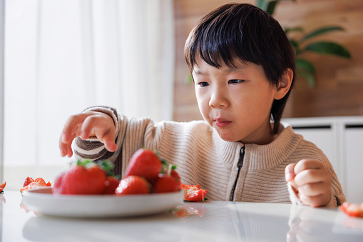 An Asian boy, about 5 years old. He is wearing a yellow sweater. Sit at the table and eat strawberries happily.