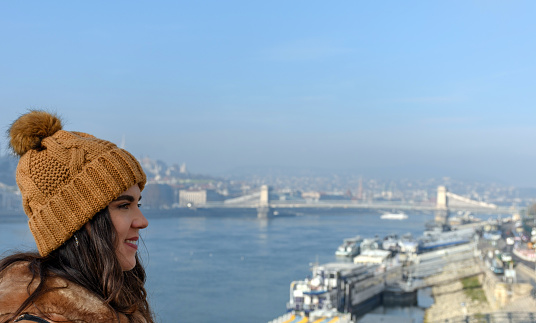A side view of a young happy Caucasian woman in winter clothes admiring Budapest and Danube river