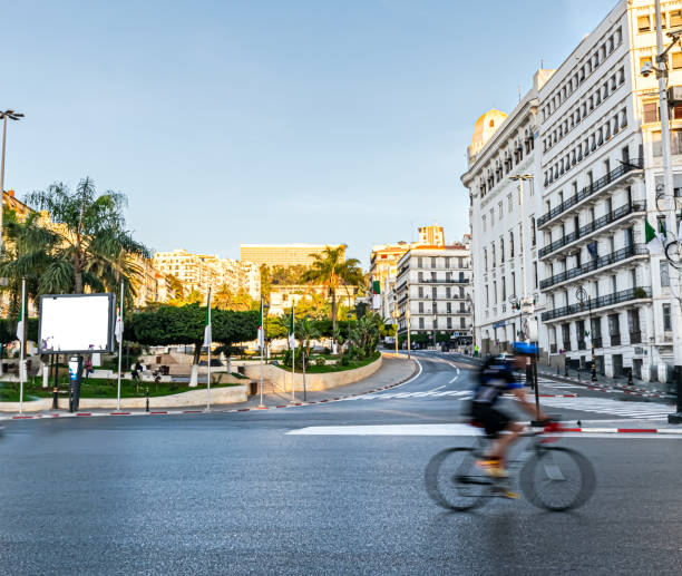 Motion blur bicycle rider in Zighoud Youcef Boulevard, Algiers city. Central post office place and building, Algerian flag posts, palm trees and the Aurassi Hotel in background with golden sunlight. algeria flag silhouettes stock pictures, royalty-free photos & images