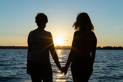 Two Girl Friends are walking along the beach holding hands on a summer day, enjoying summer holidays. Silhouette of two women at sunset.