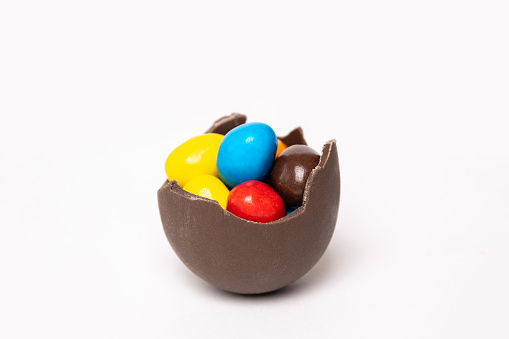 Cracked chocolate easter egg with colorful small round candies on white background, copy space, side view. Chocolate treat for kids. Easter concept.