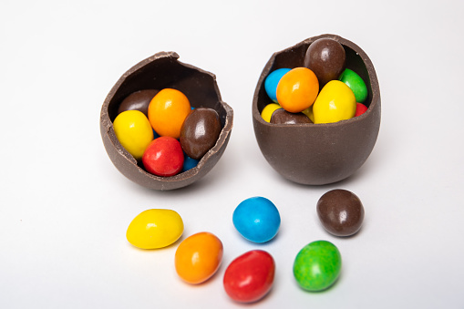 Cracked chocolate easter eggs full of colorful small round candies on a white background with colorful candies, copy space, top view. Chocolate treat for kids. Easter concept.