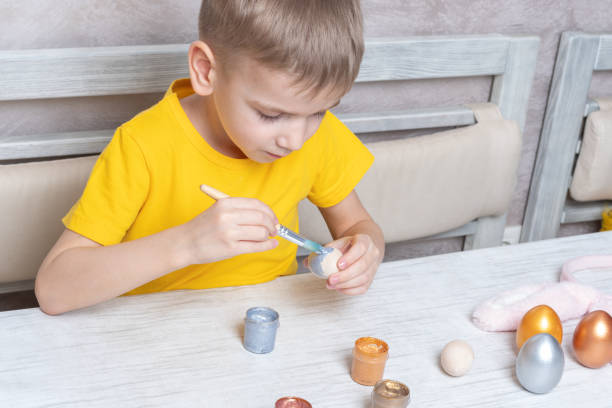 um garotinho loiro pinta ovos para o feriado de páscoa em casa. a criança se diverte e comemora o feriado - face paint child paint rabbit - fotografias e filmes do acervo