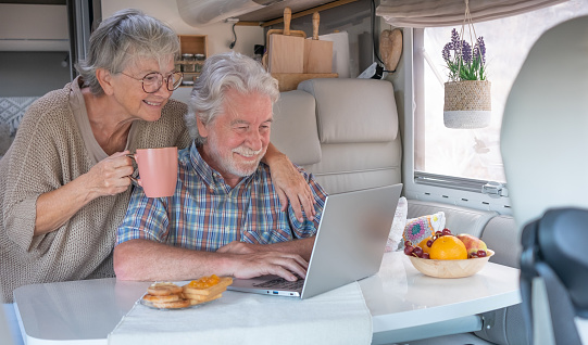 Beautiful happy caucasian senior couple in travel vacation leisure sitting inside a camper van dinette enjoying breakfast together while using laptop