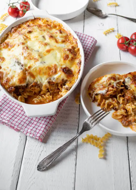 Homemade pasta casserole, italian cuisine with fusilli noodles, bolognese sauce, bechamel sauce and mozzarella cheese. Served open in a casserole dish with plates on white wooden background with decoration. Top view with space for text
