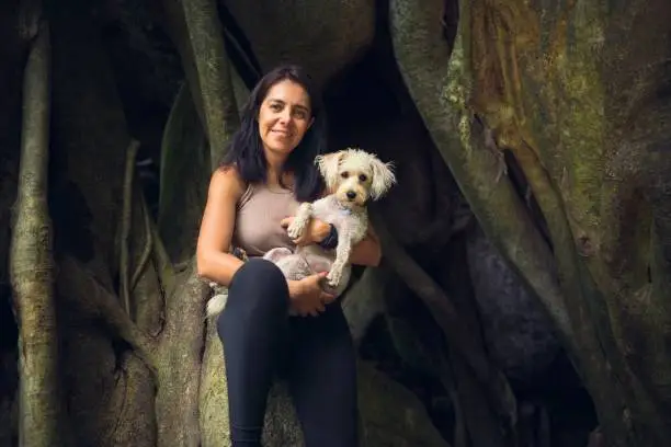 A smiling Hispanic woman with an adorable white Schnoodle in her arms against a giant jungle tree