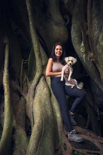 A vertical view of a smiling Hispanic woman holding a white Schnoodle against a giant jungle tree