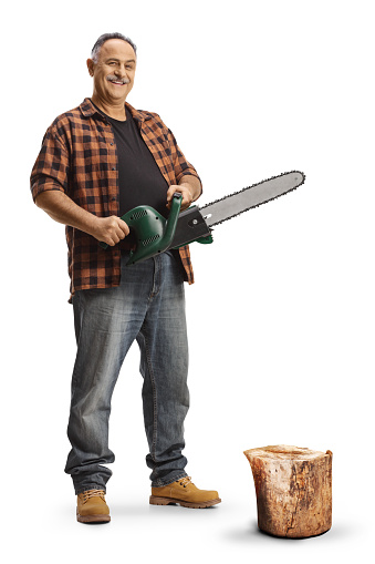 Full length portrait of a mature man with a chainsaw standing next to a tree trunk isolated on blue background