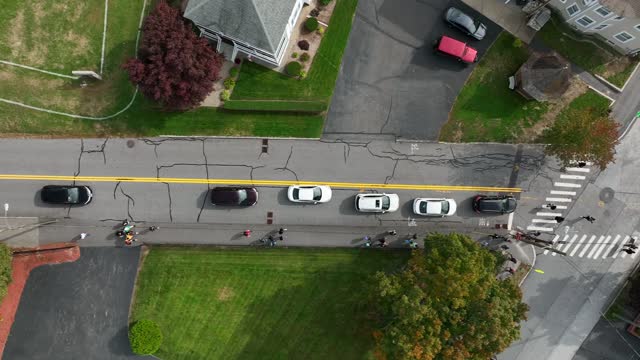 Straight down aerial shot of sedan cars driving down street and coming to stop at intersection where pedestrians cross crosswalk. Fall foliage lines sidewalks and streets where locals travel.