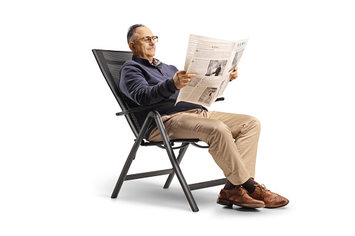 Casual mature man sitting in a foldable chair and reading a newspaper isolated on white background