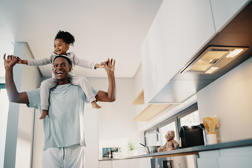 Father carrying his daughter on his shoulders, he smiles and holds her hands outstretched. Man playing with his daughter at home. Family fun between father and daughter.