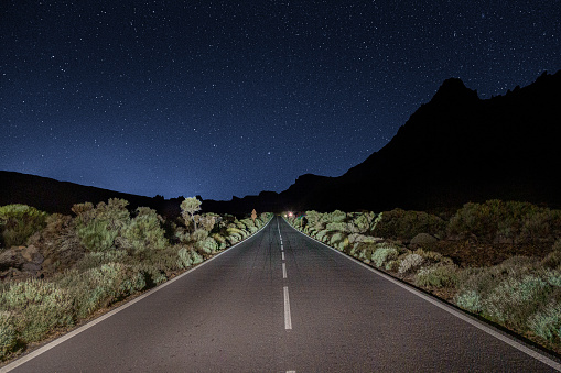 Brightly lit straight highway with green bushes on both sids, with a silhouette of the tall rocks and the dark blue night sky and subtle set of stars