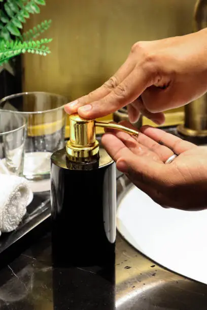 Photo of Close-up image unrecognisable person wearing bathrobe using black pump soap dispenser for hand washing, artificial fern plant on black marble bathroom counter, white bathroom sink, drinking glass, rolled flannels, focus on foreground