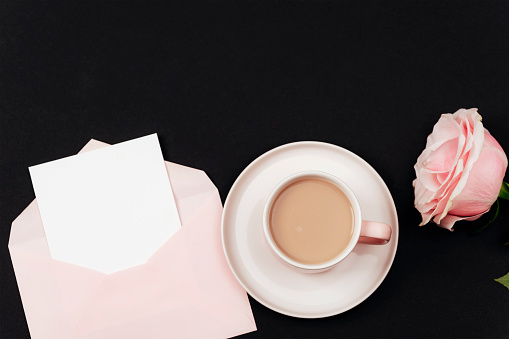 Coffee cup, envelope with blank card and pink rose on black background. Top view, flat lay, copy space.